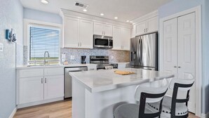 Een moderne keuken met witte kastjes, roestvrijstalen apparatuur, een groot eiland en een raam met jaloezieën.