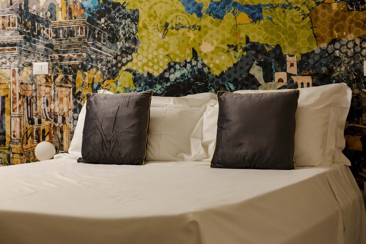 Un lit avec une literie blanche et deux oreillers foncés, placé devant un mur décoré d’une fresque abstraite aux couleurs vives.