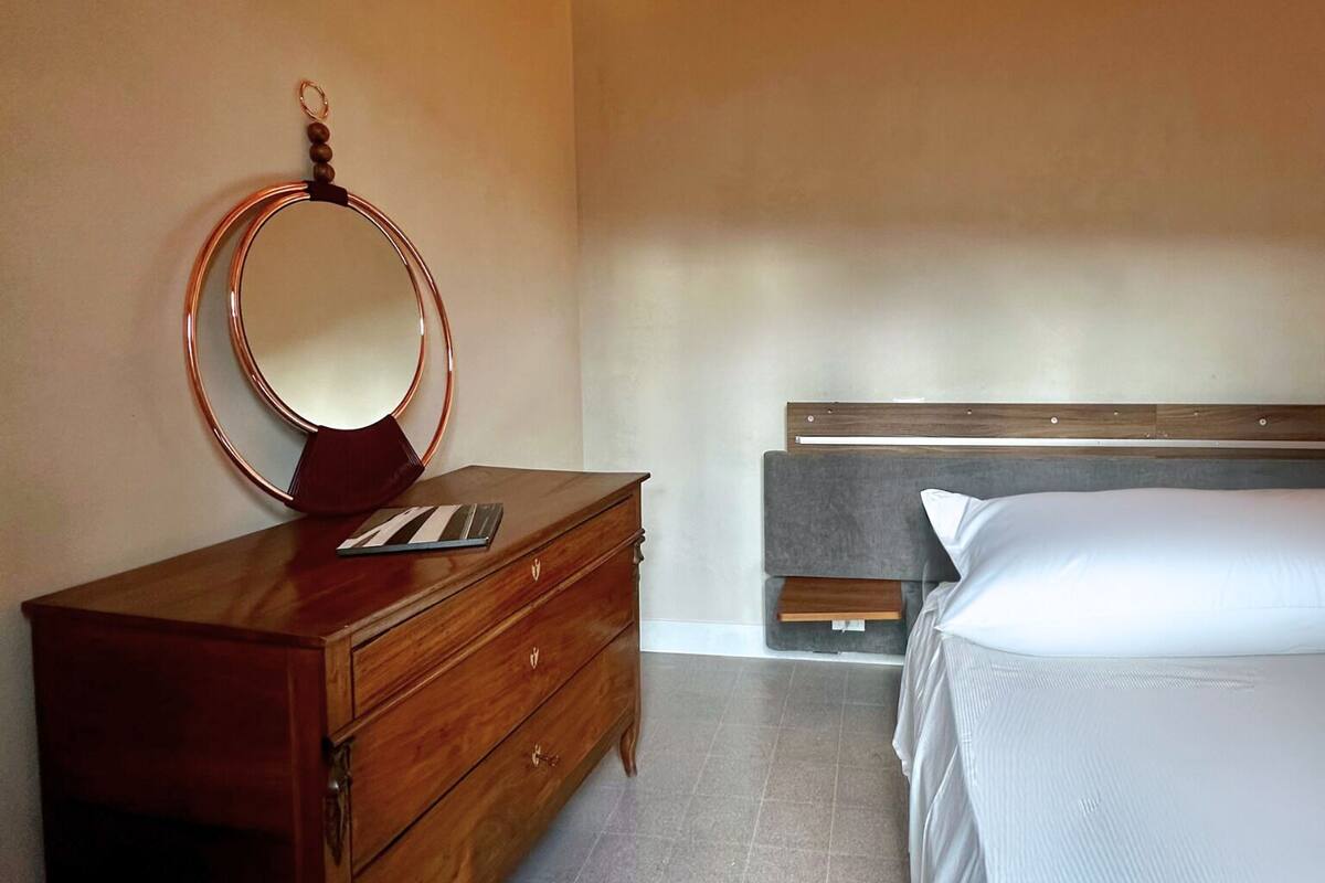 Ein Schlafzimmer mit einem hölzernen Schminktisch, einem runden Spiegel, einem Bett mit grauem Kopfteil und einer schrägen Decke mit freiliegenden Balken.