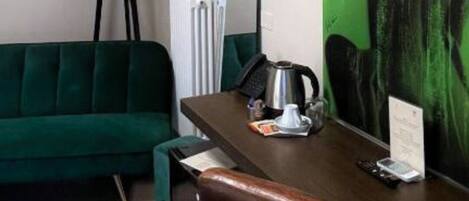 Habitación de hotel con un sofá verde, un escritorio con cafetera, una lámpara y un cuadro grande de color verde en la pared.