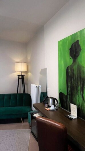 Une chambre d’hôtel avec un canapé vert, un bureau avec une machine à café, une lampe et un grand tableau vert accroché au mur.
