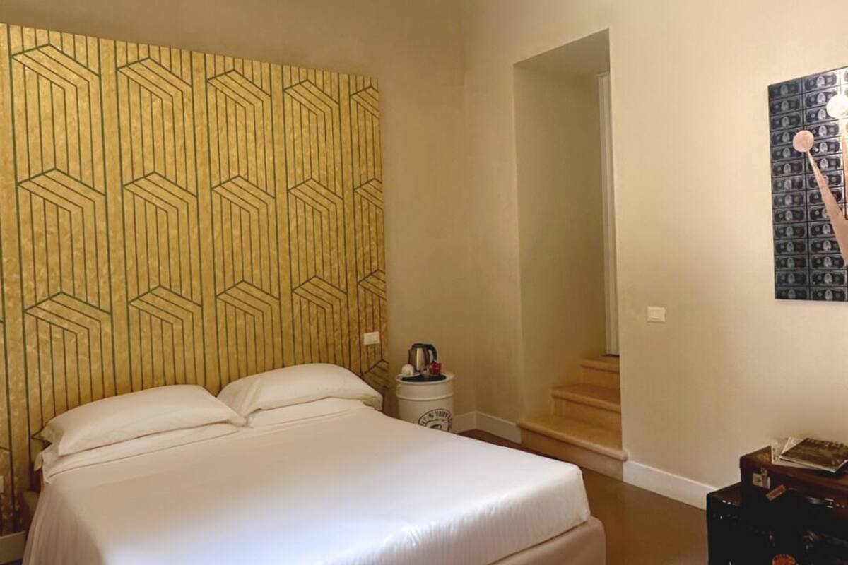 Ein Schlafzimmer mit einem Bett, einer Wand mit geometrischem Muster, einer Deckenleuchte, einem Nachttisch, einem Koffer und einem gerahmten Bild.