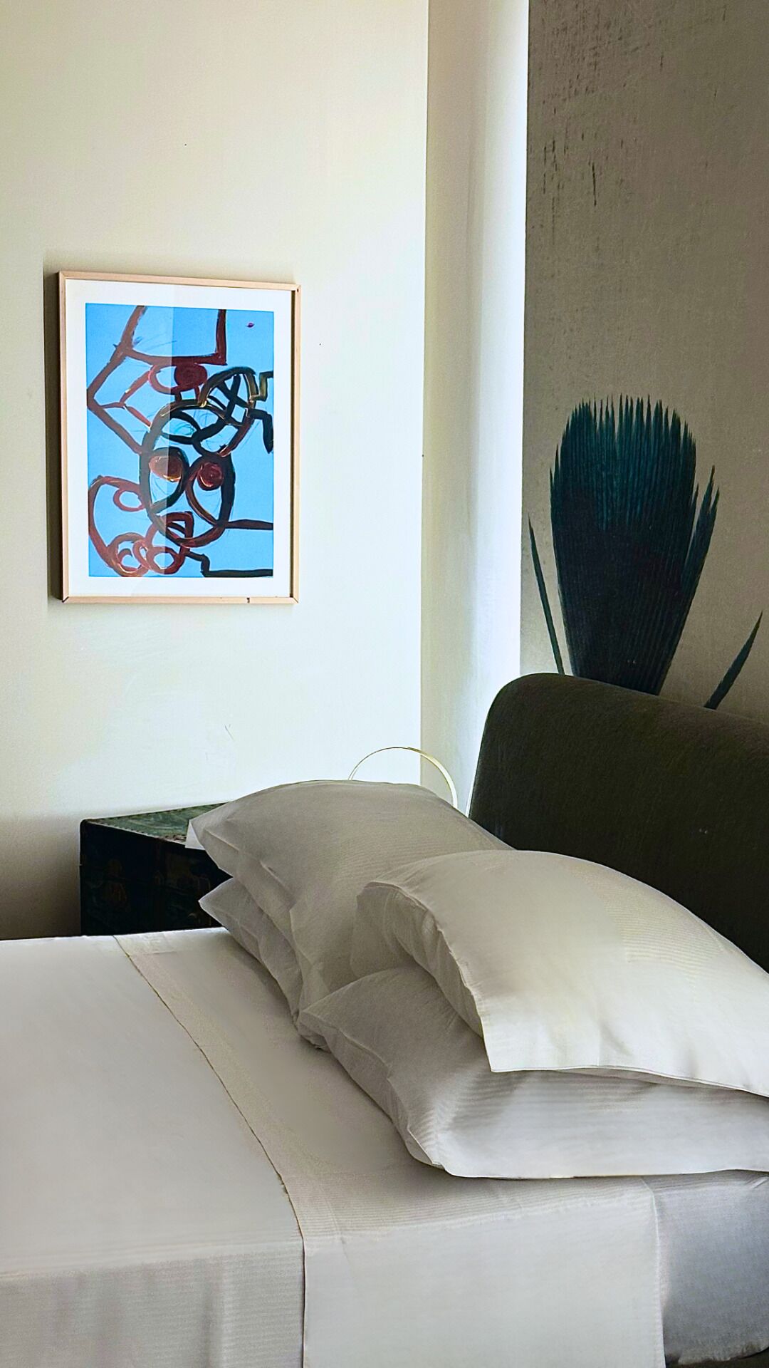 Un letto rifatto con lenzuola e cuscini bianchi, un quadro astratto incorniciato appeso alla parete e una sedia scura dal design originale.