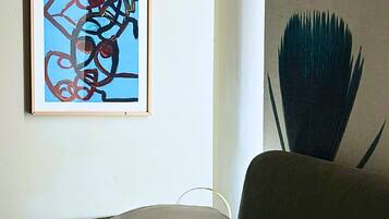 Un letto rifatto con lenzuola e cuscini bianchi, un quadro astratto incorniciato appeso alla parete e una sedia scura dal design originale.