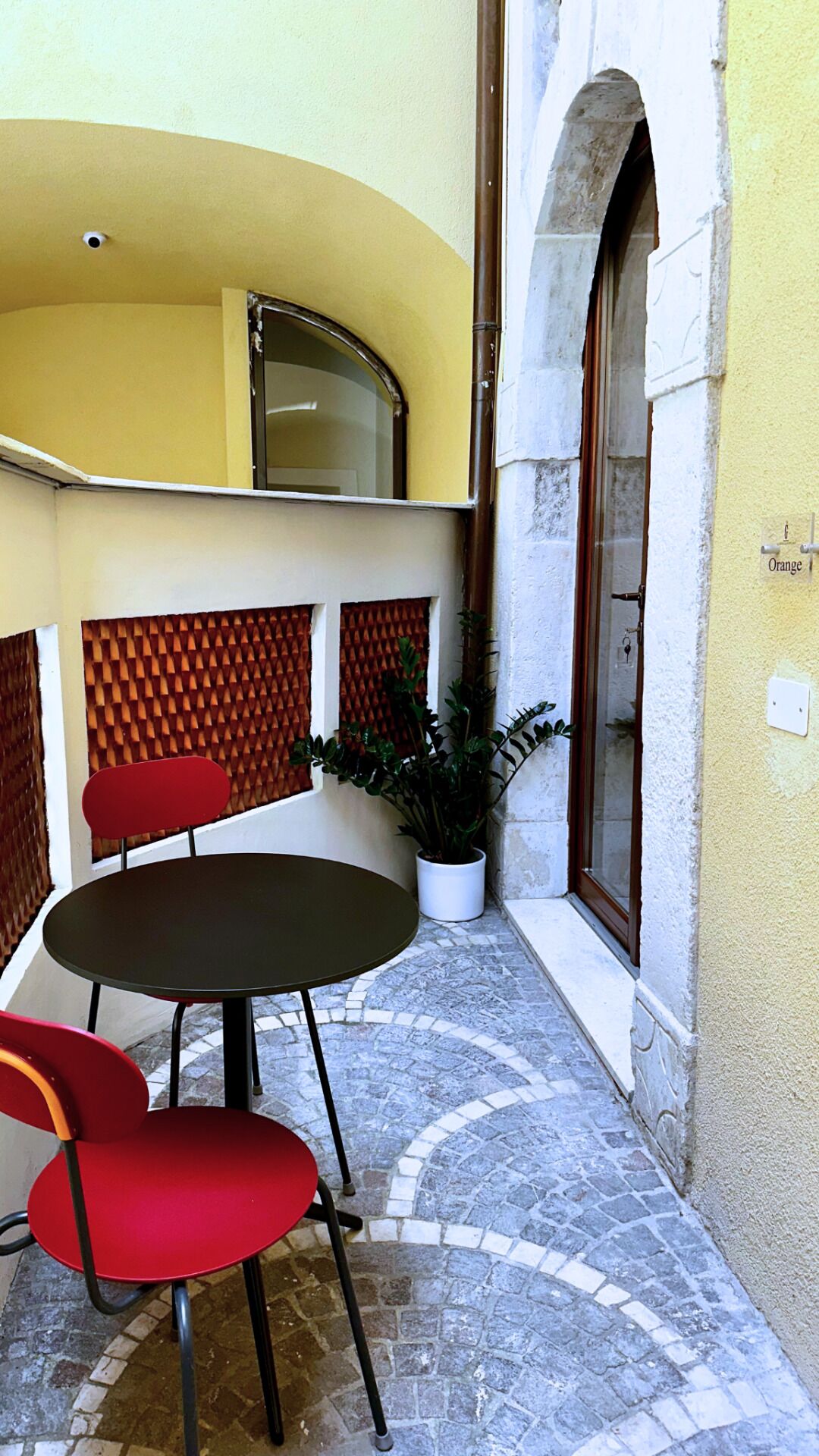 A small outdoor seating area with a round table and two red chairs, a potted plant, and a stone-tiled floor.