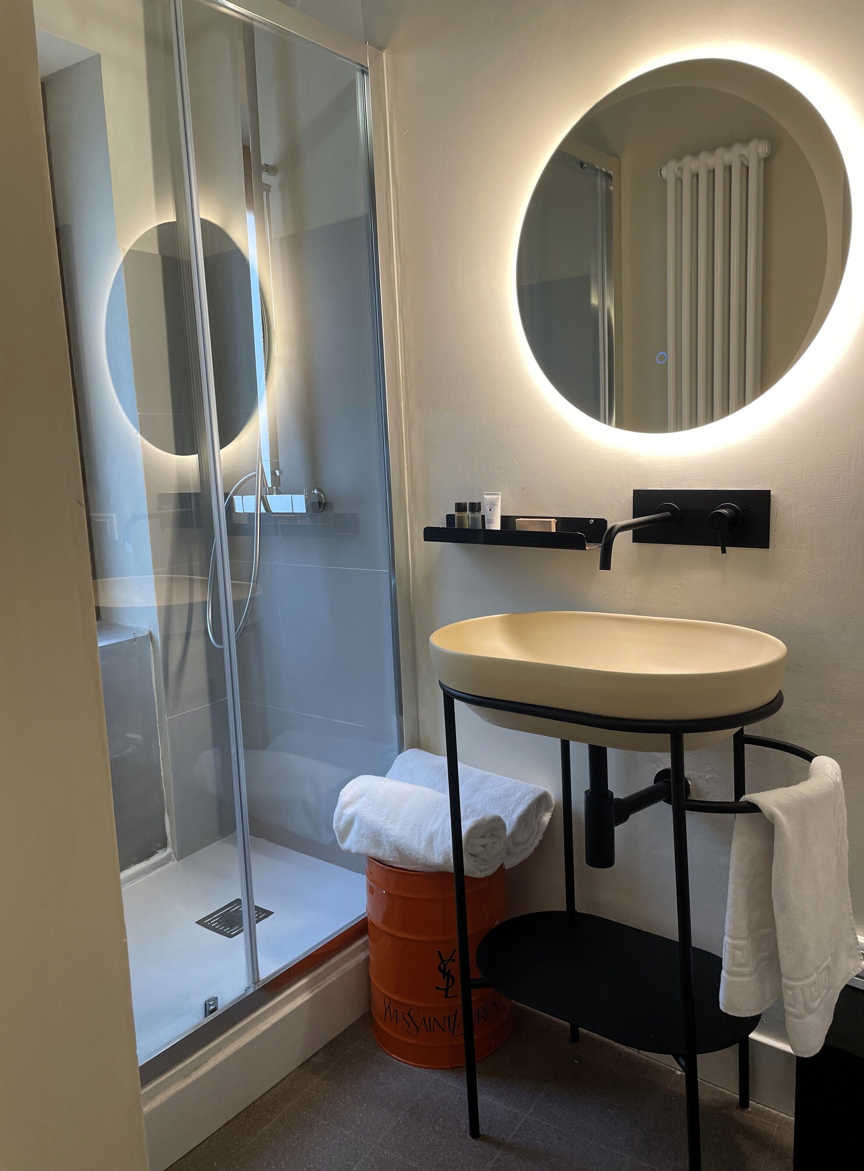 Un baño moderno con cabina de ducha de vidrio, un espejo redondo, un lavamanos con un taburete negro y un barril naranja grande.