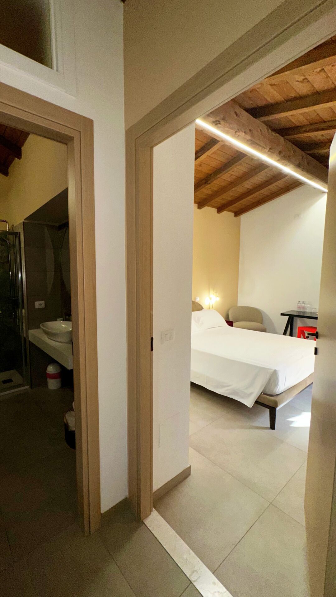 A bedroom with a bed, a bedside table, and a nightstand. There is a bathroom with a sink and a toilet visible through an open door.