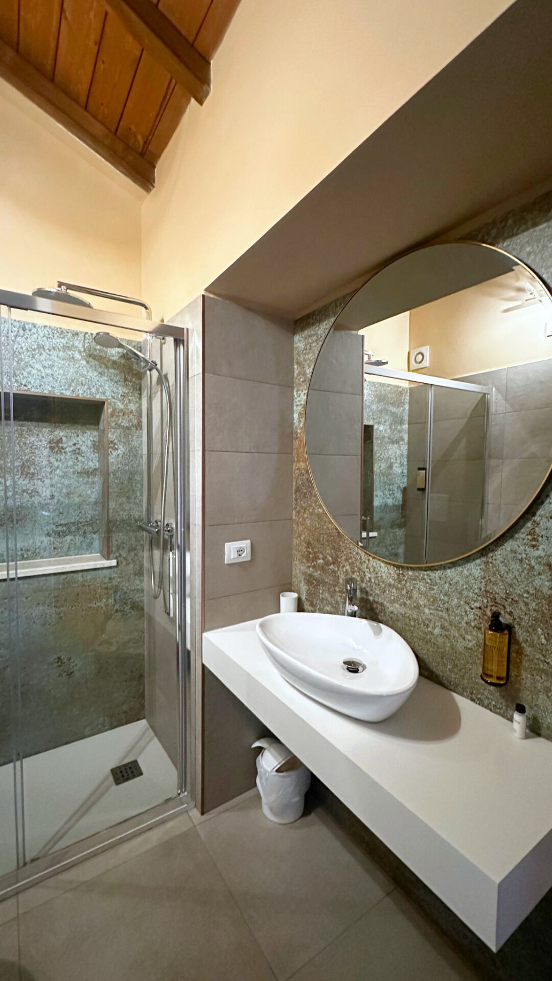 A modern bathroom with a glass shower enclosure, a white sink with a marble backsplash, and a large round mirror.
