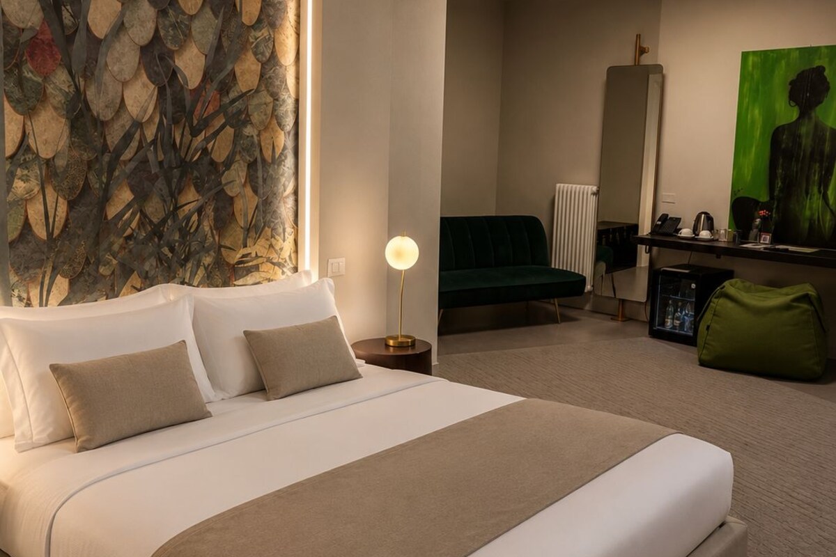 Une chambre d’hôtel moderne avec un grand lit, un mur décoratif en pierre, un canapé vert, un bureau avec une lampe et un tableau.