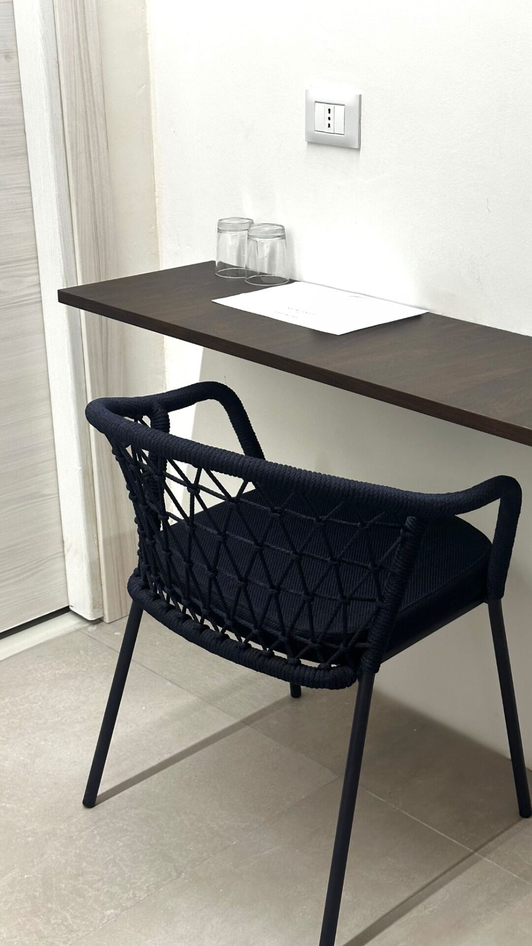 Uma mesa de madeira escura com uma tomada na parede branca, dois copos de vidro transparentes e uma cadeira de fibra preta.