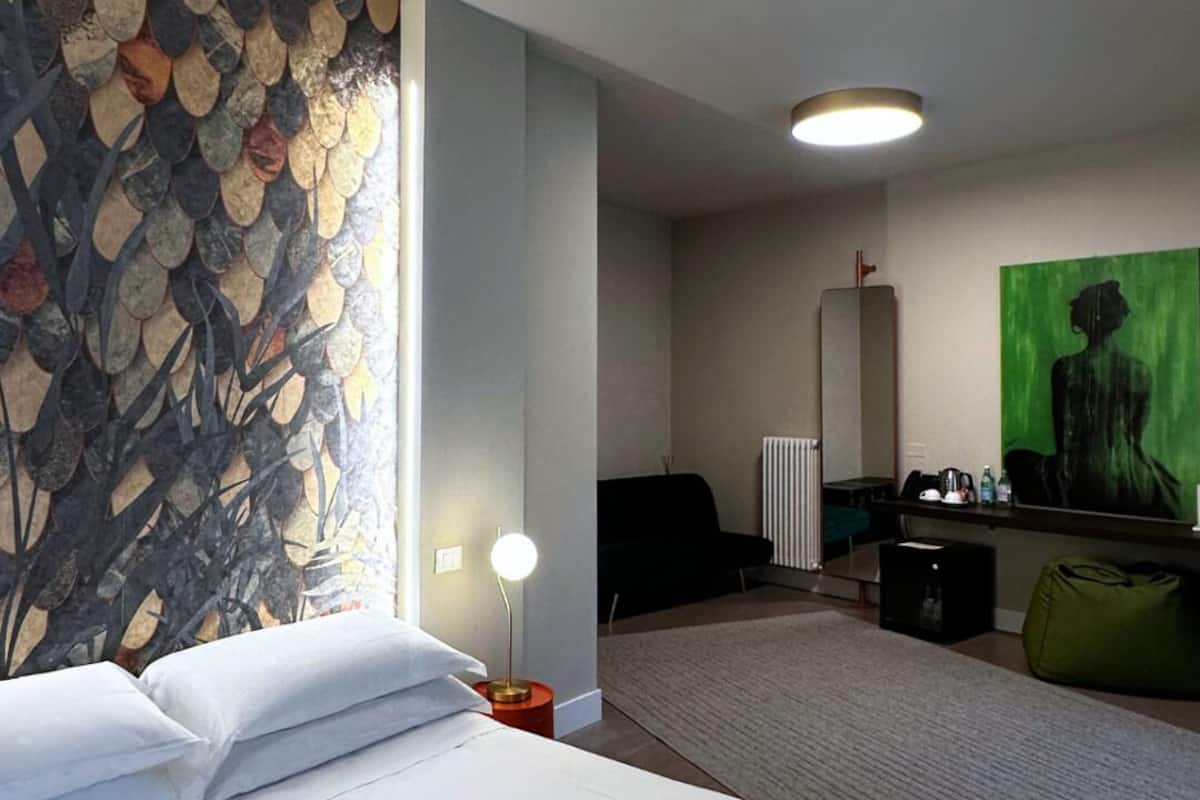 Ein Hotelzimmer mit einem großen Bett, einem Wandbild, einem Heizkörper, einem Schreibtisch und einem grünen Sessel.
