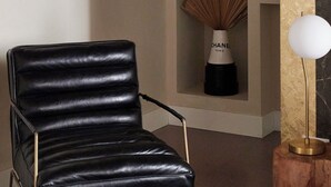 Een zwarte leren fauteuil met metalen poten, een klein houten bijzettafeltje met een witte lamp en een decoratief object met een Chanel-label op de achtergrond.