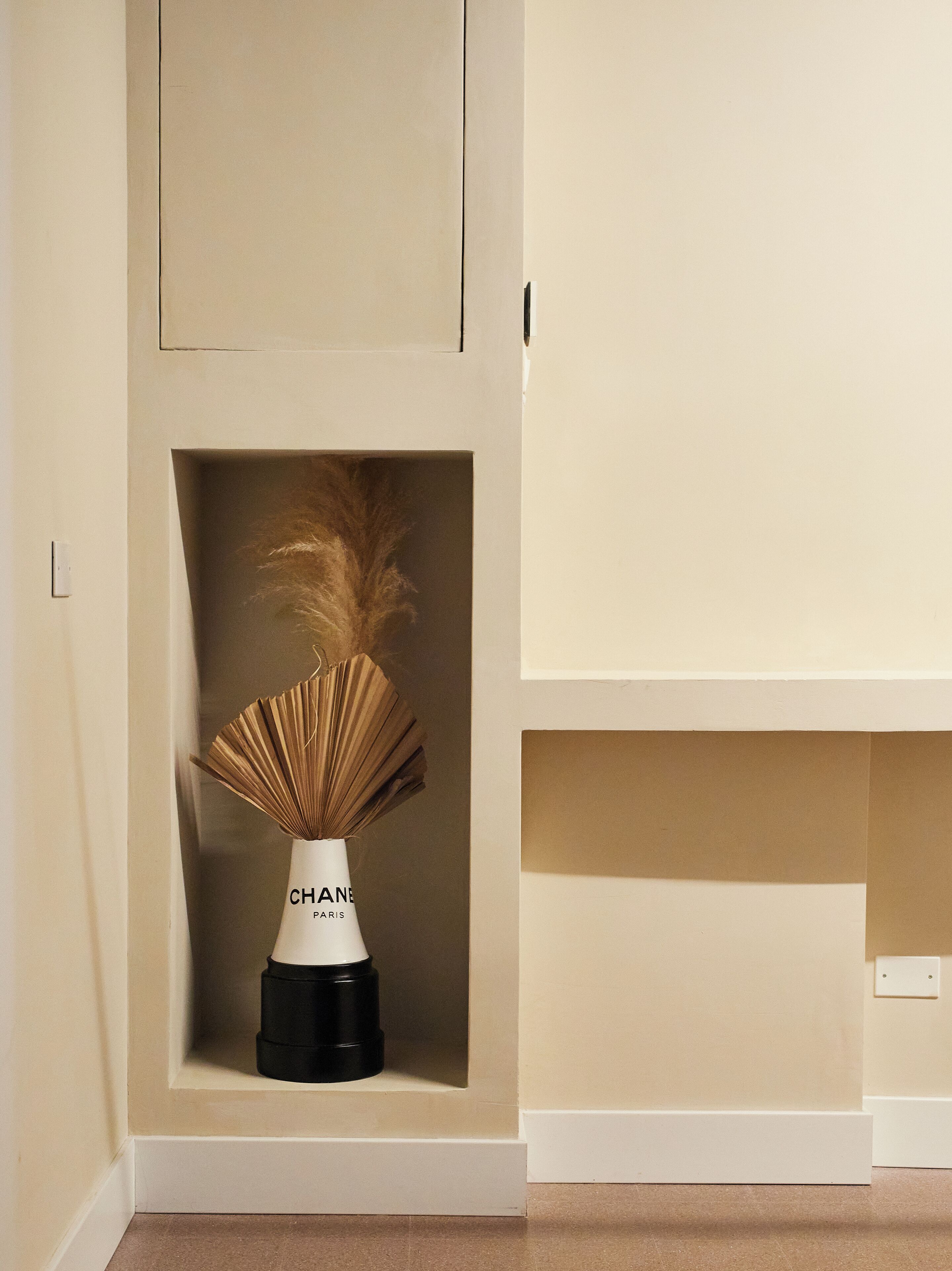 Un vase blanc avec une base noire portant l’inscription « CHANEL PARIS », le tout placé sur un mur de couleur beige.