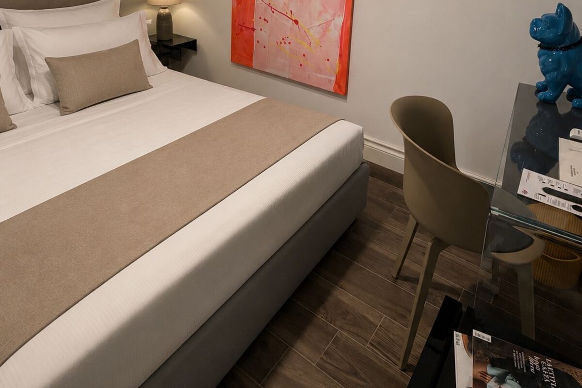Une chambre d’hôtel moderne avec un grand lit, une table de chevet avec une lampe, un tableau accroché au mur, un porte-manteau suspendu et une figurine de chien bleue posée sur une table d’appoint.