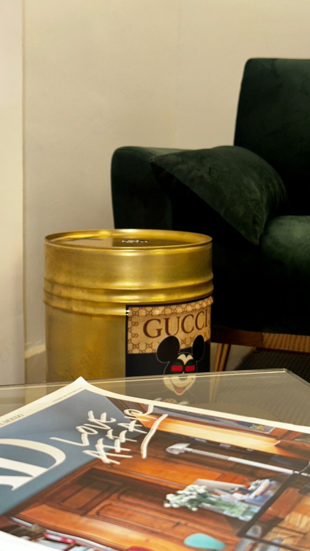 A glass table with a magazine, a gold-colored container with a Gucci label, and a dark green chair.