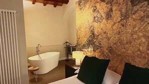 Een slaapkamer met een groot bed, een houten plafond, een stenen muur, een witte badkuip en een klein krukje.