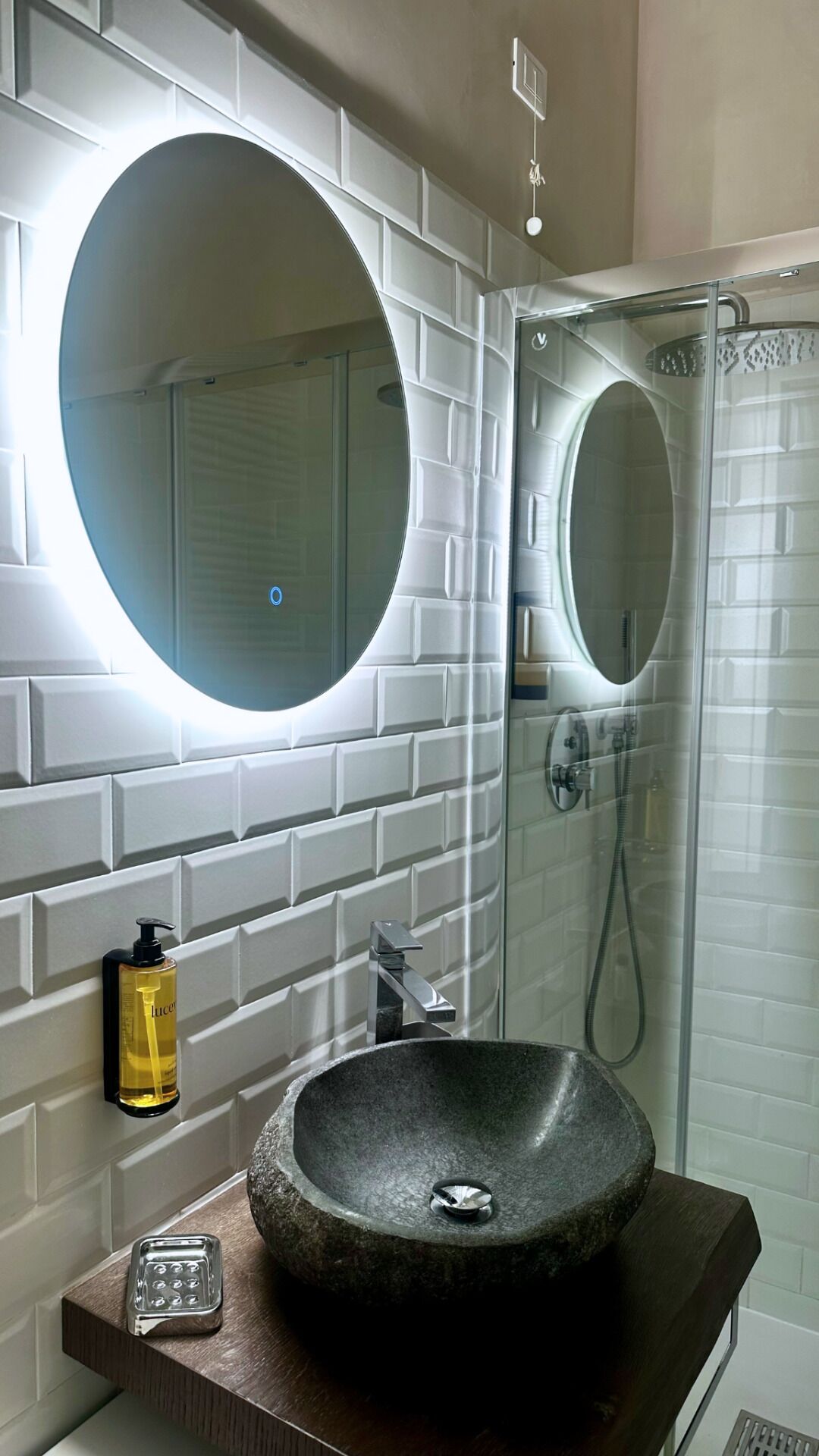 Un bagno con uno specchio rotondo, un lavabo con una vasca in pietra e un box doccia in vetro.