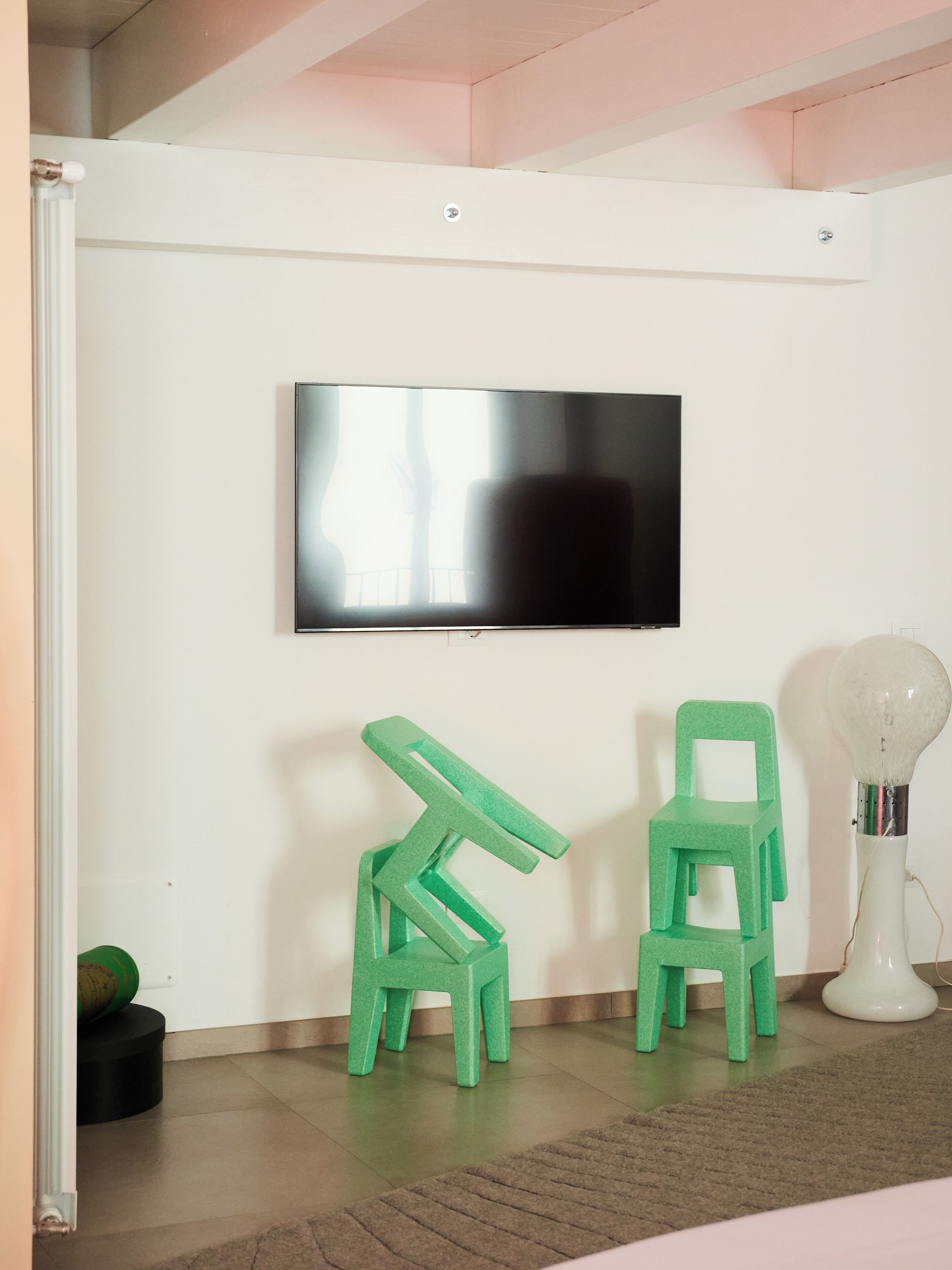 A room with a flat-screen TV mounted on the wall, two green plastic chairs, a small green plant, and a white lamp on the floor.