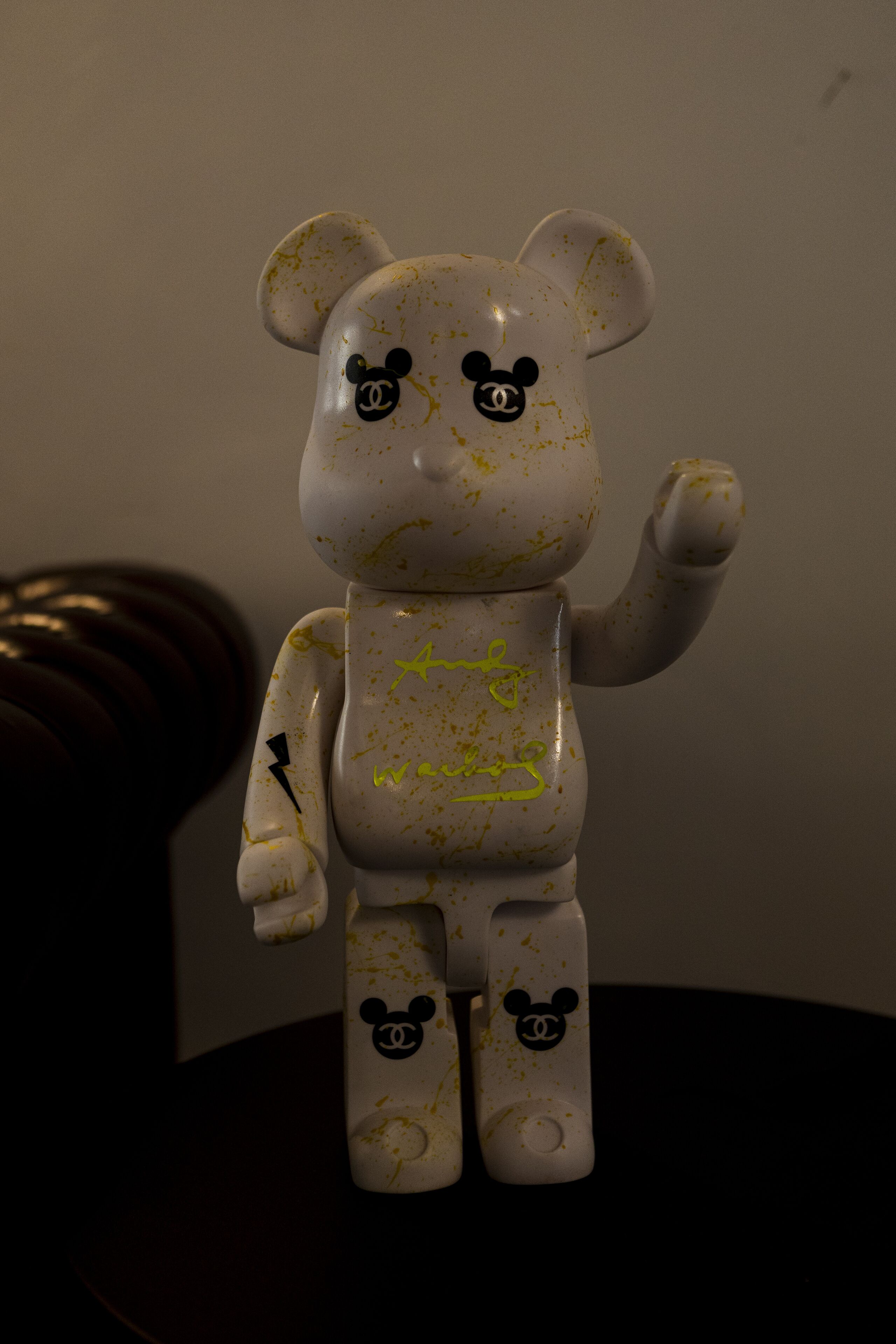 En hvid bamsefigur med gule stænk og sorte Mickey Mouse-logoer på kroppen, stående på en mørk overflade.