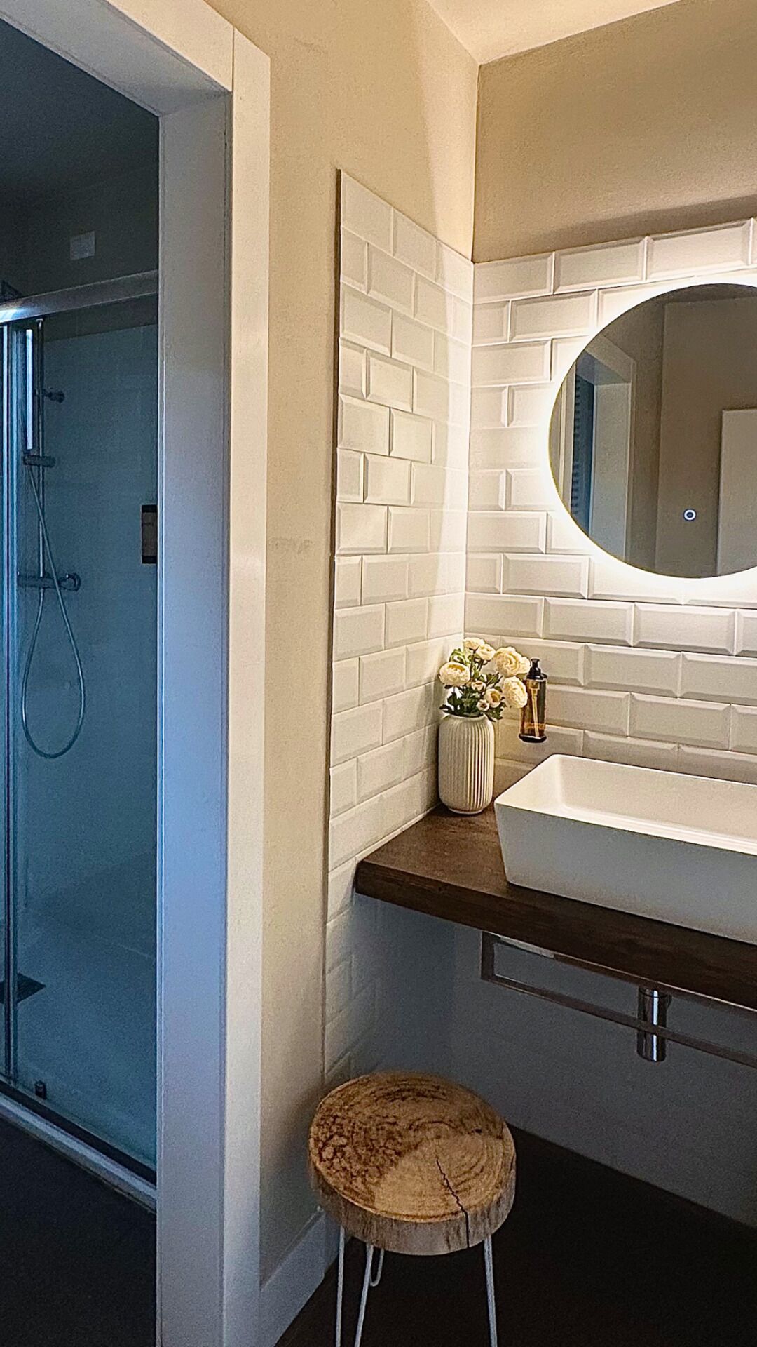 A bathroom with a glass shower enclosure, a white tiled wall, a wooden stool, and a round mirror above a sink.