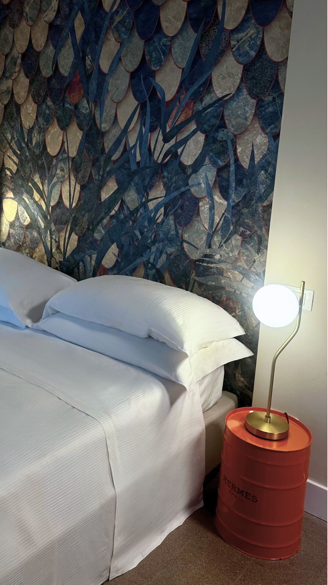 A bed with white linens, a red barrel lamp, and a wall with a blue and beige abstract pattern.