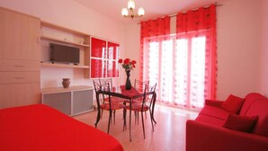 Une pièce comprenant une table à manger, des chaises, un téléviseur posé sur une étagère, un canapé rouge et une fenêtre avec des rideaux rouges.