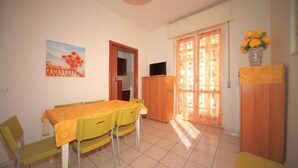 Uma sala de jantar com mesa e cadeiras, uma televisão em um móvel, e uma porta com cortinas.