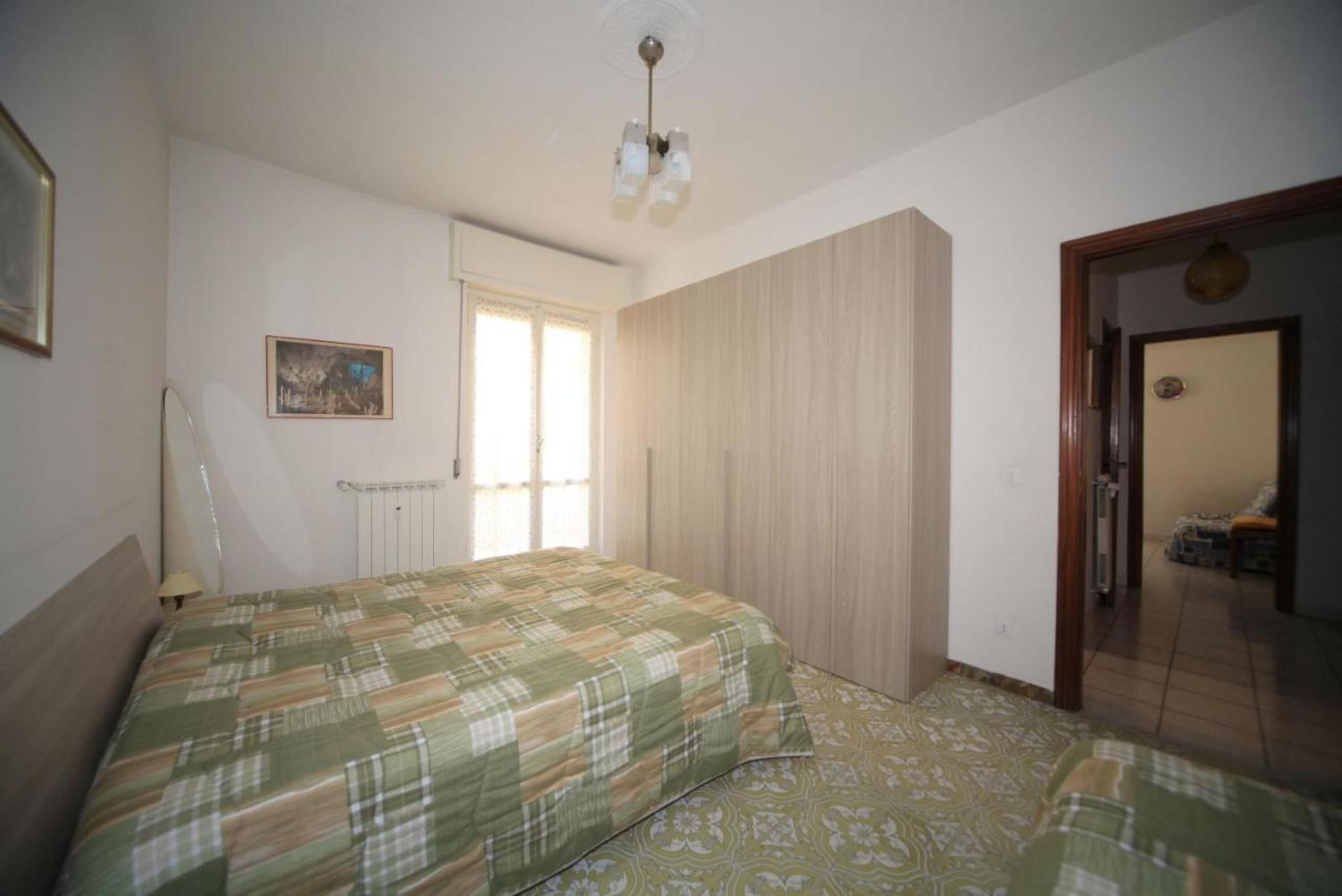 A bedroom with a bed, a radiator, a window, and a door leading to another room.