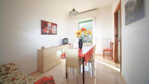 A bright living room with a sofa, a dining table with chairs, a TV, and a vase with flowers.