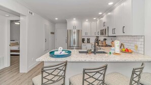 A modern kitchen with white cabinets, a stainless steel refrigerator, a sink, and a bar area with stools. - Ocean Forest Villas A-305 (Myrtle Beach)