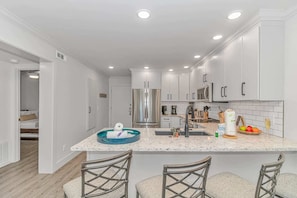 A modern kitchen with white cabinets, a stainless steel refrigerator, a sink, and a bar area with stools.