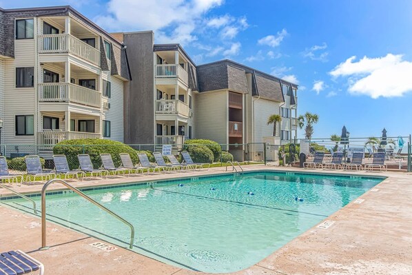 A swimming pool with lounge chairs, a lifeguard sign, and a multi-story building with balconies.
