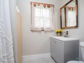 A bathroom with a white sink, a mirror, and a window with a curtain. - Cottage #6 - Cute and airy 2 bedroom cottage right on Chautauqua Lake! (Ashville)
