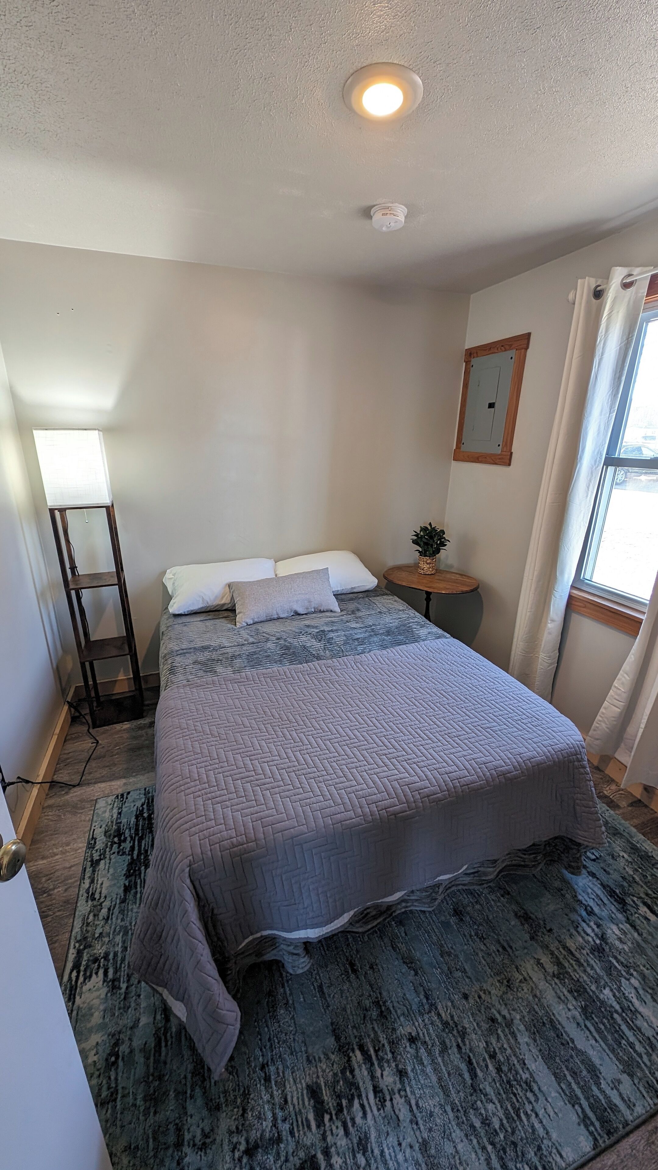 Une chambre à coucher comprenant un lit, une table de chevet, une lampe, une fenêtre avec des rideaux et un luminaire fixé au mur.