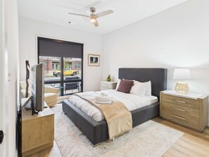 Une chambre moderne comprenant un lit, des tables de chevet, un bureau, une chaise, un ventilateur de plafond et une fenêtre donnant sur l’extérieur.