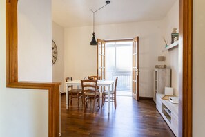 A dining area with a table and chairs, a clock on the wall, and a window with an open door leading outside.