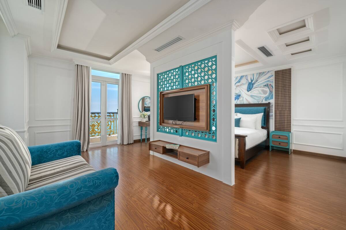 Ein geräumiges Hotelzimmer mit einem großen Bett, einem an einer Zierwand montierten Fernseher, einer Couch und einem Balkon mit Vorhängen.