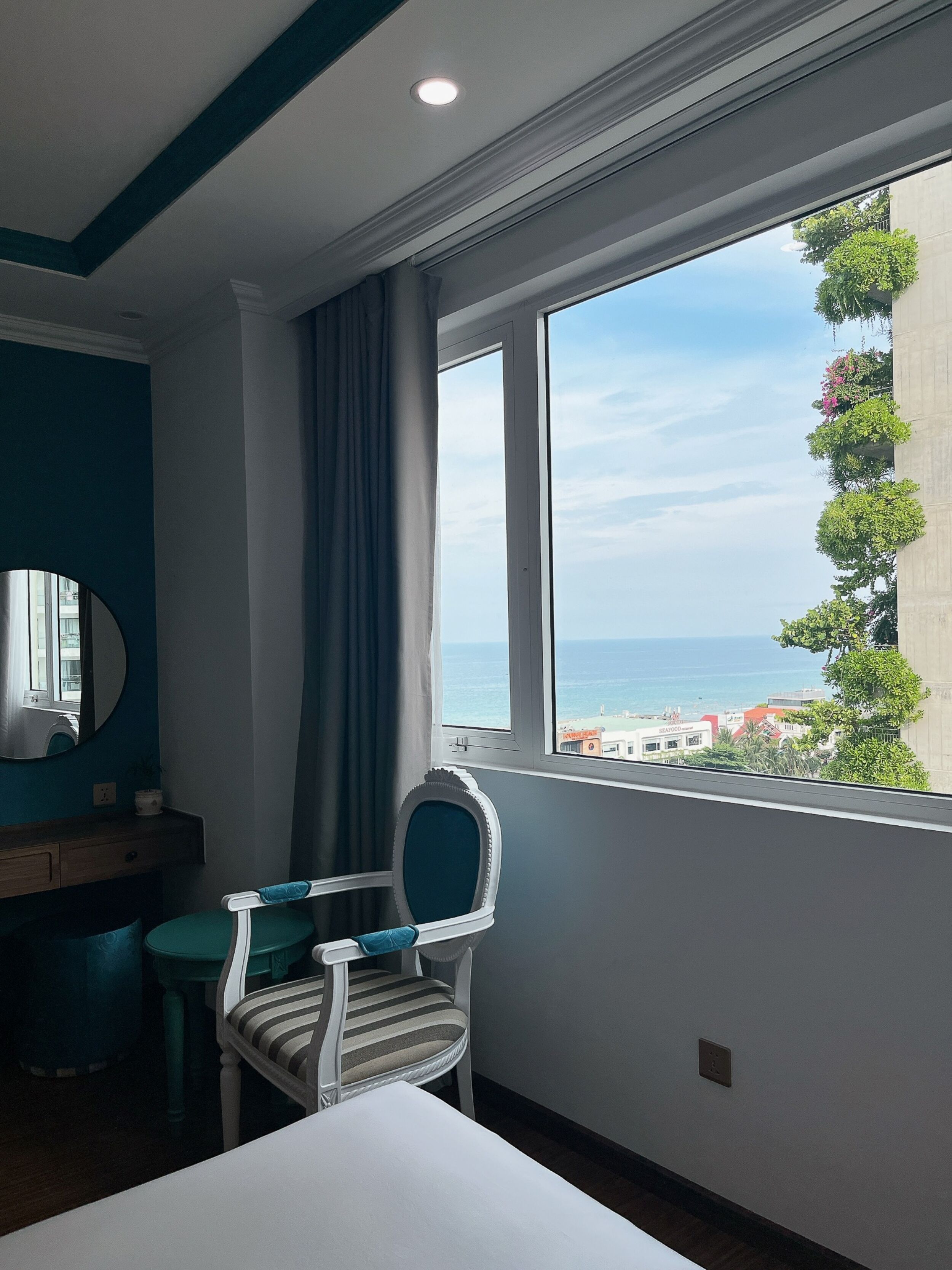 A hotel room with a large window offering a view of the sea and buildings.