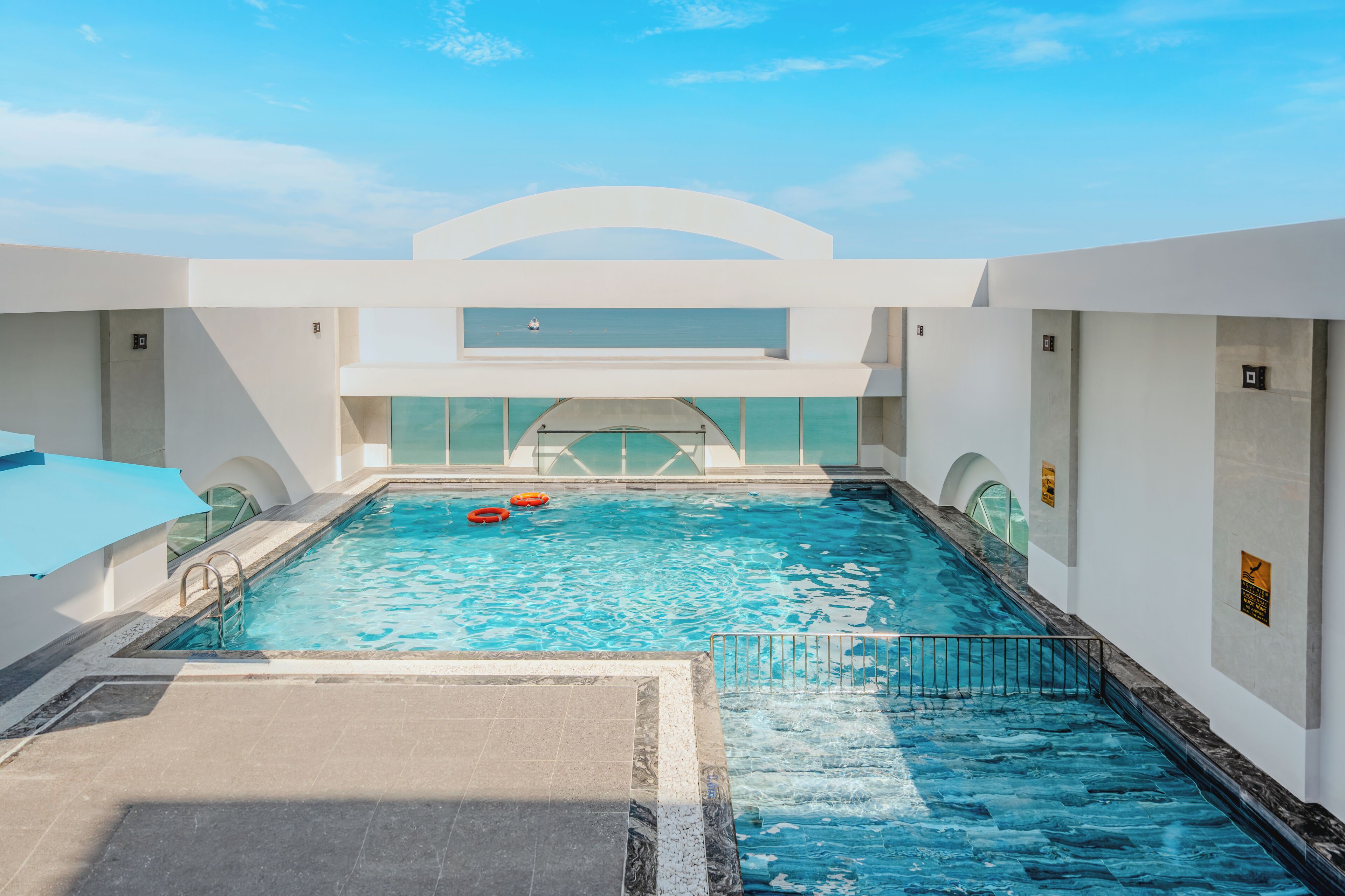Una piscina interior moderna con agua azul cristalina, una sombrilla azul y vistas al mar a través de un amplio ventanal.