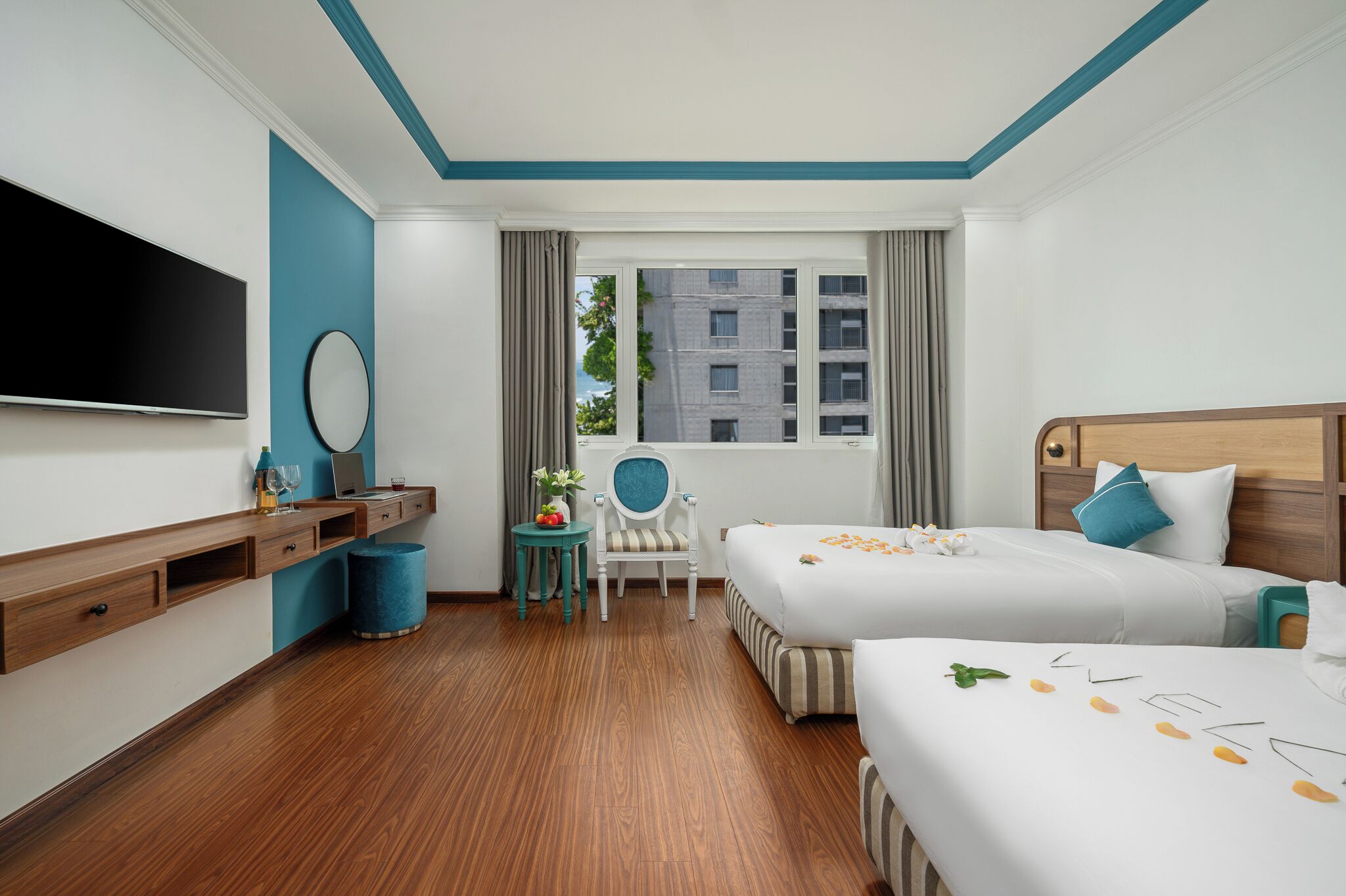 Habitación de hotel con dos camas, televisión, escritorio con silla, ventana con vistas a edificios y una pared de acento azul.