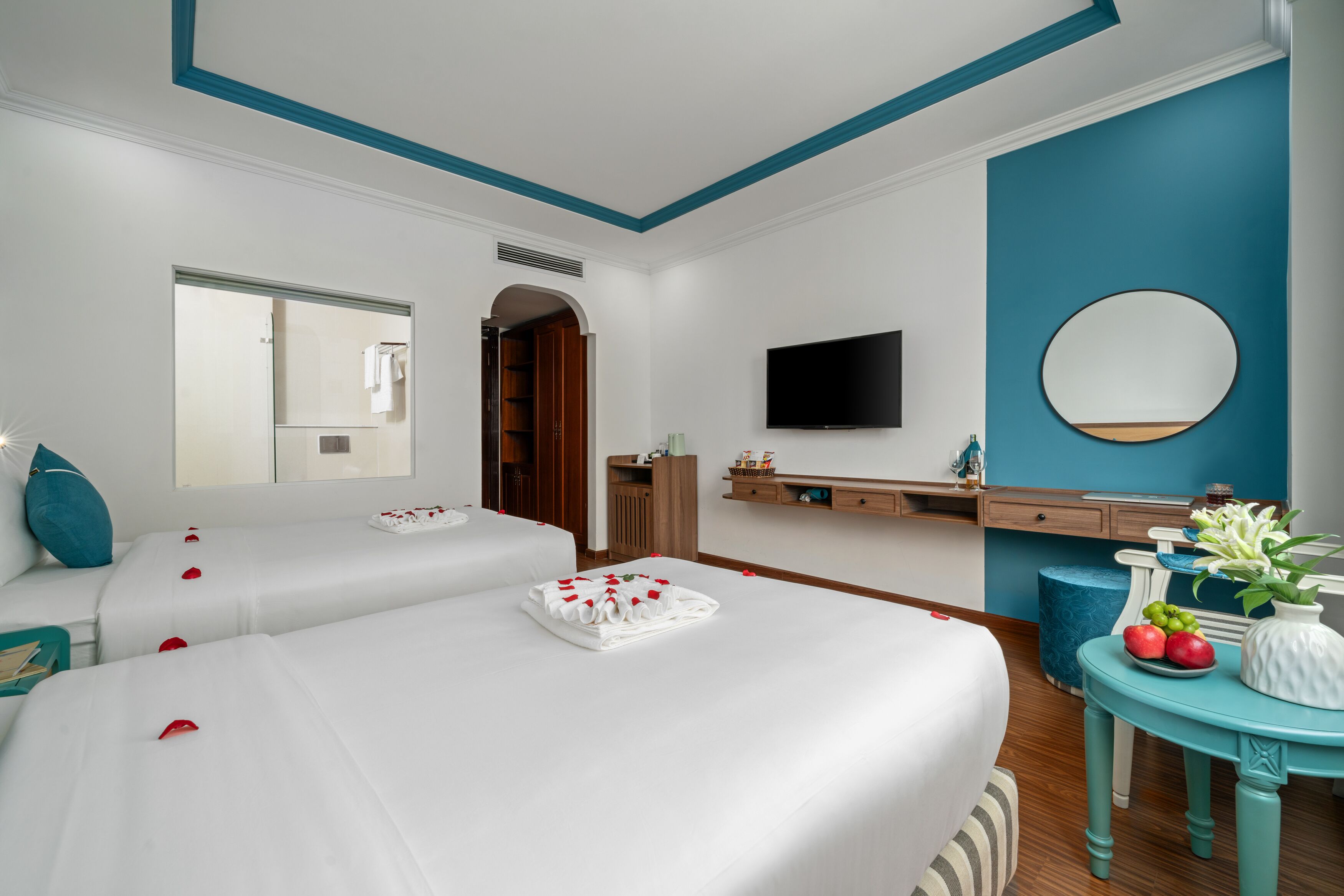Quarto de hotel com duas camas, uma TV, um espelho, toucador com pia e uma pequena mesa com frutas e flores.