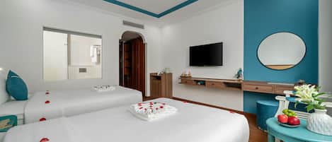 Quarto de hotel com duas camas, uma TV, um espelho, toucador com pia e uma pequena mesa com frutas e flores.