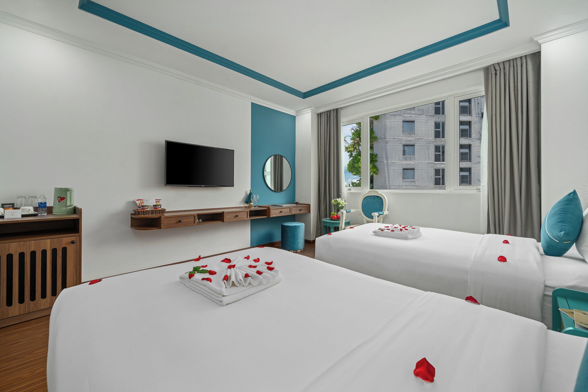A hotel room with two beds, a TV, a mirror, a window with curtains, and a view of buildings.