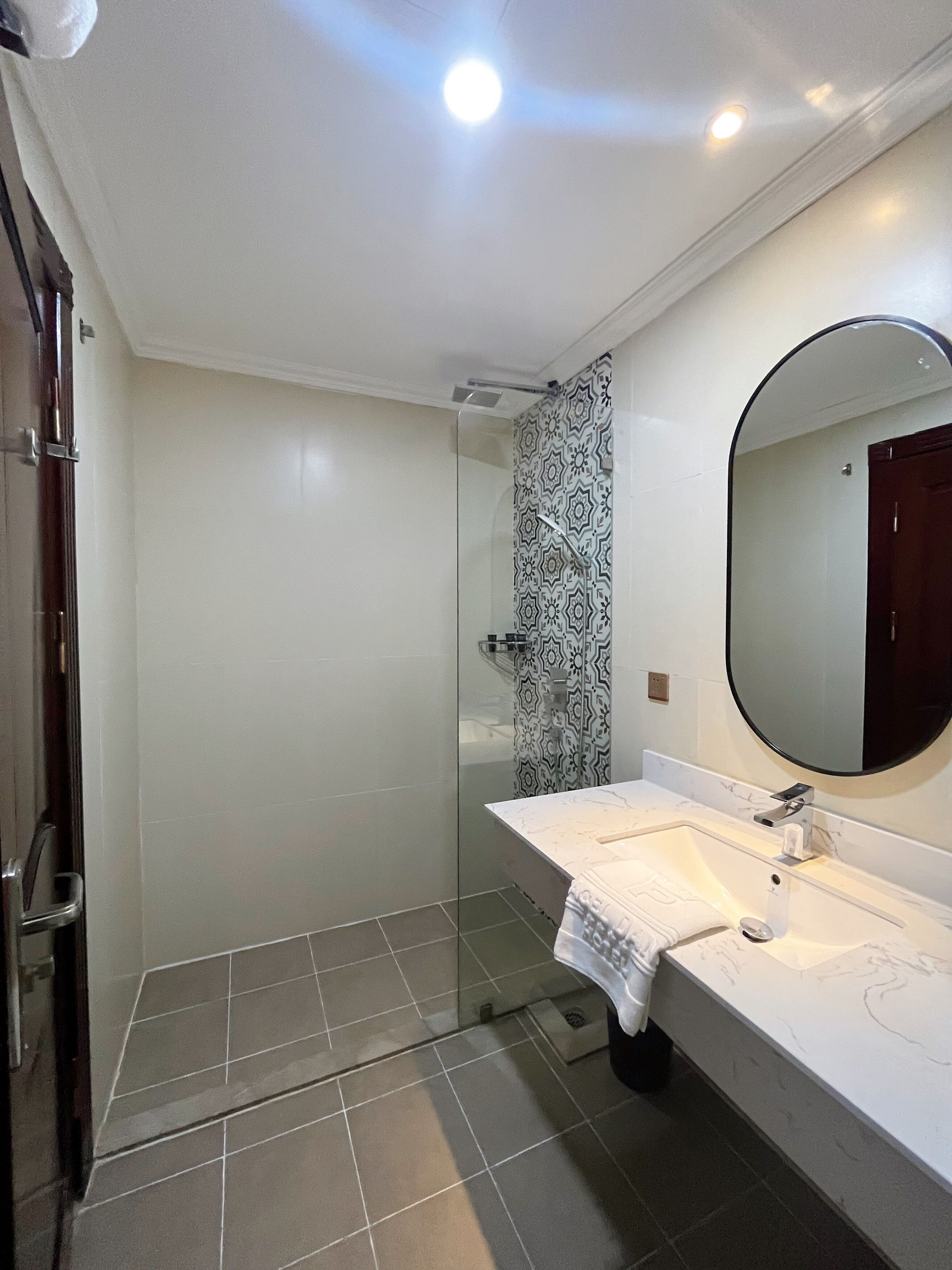 A bathroom with a glass shower enclosure, a white sink with a marble countertop, and a round mirror above the sink.