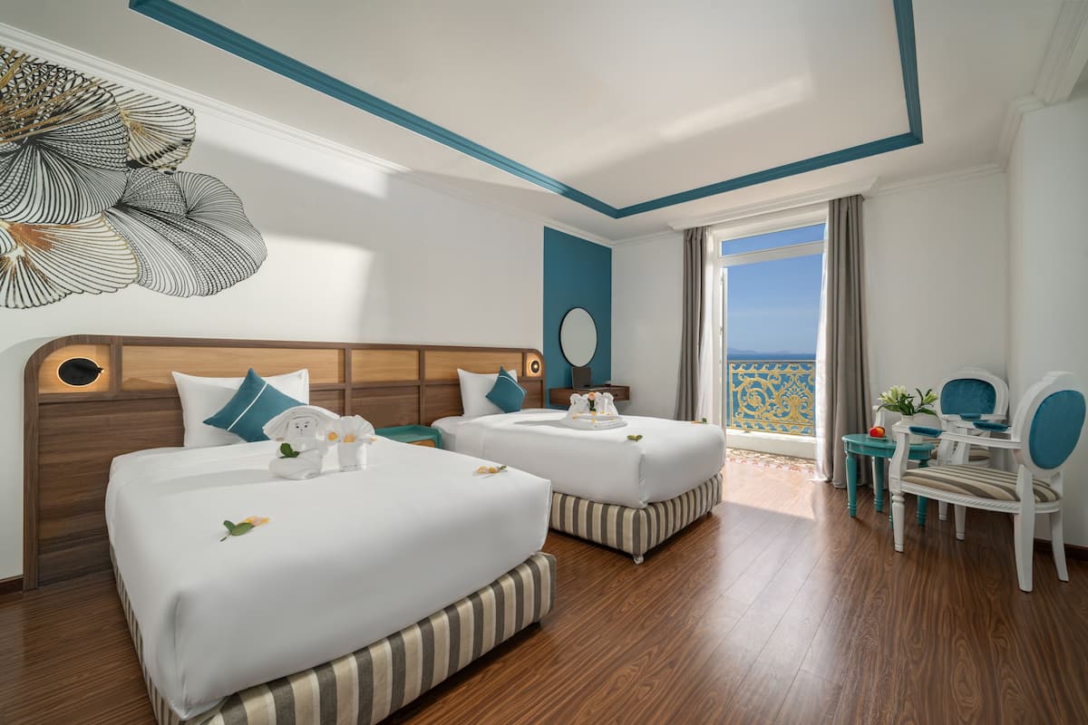 Ein Hotelzimmer mit zwei Betten, einem Holz-Kopfende, einem runden Spiegel, einem kleinen Tisch mit einer Pflanze und Blick auf das Meer.