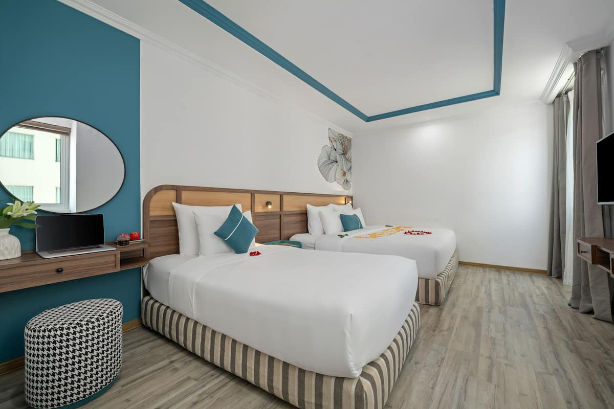 Ein Hotelzimmer mit zwei Betten, einem Holzkopfteil, einem Spiegel, einem Schreibtisch mit einem Laptop und einem Fenster mit Vorhängen.