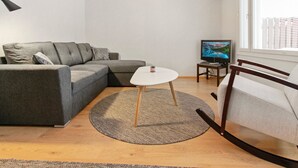 Un salon moderne avec un canapé d’angle gris, une table basse ronde, un téléviseur posé sur un meuble et une grande fenêtre équipée de stores.