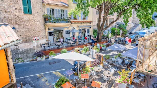 Ein Straßencafé mit Tischen und Stühlen, umgeben von Gebäuden und einem Baum.