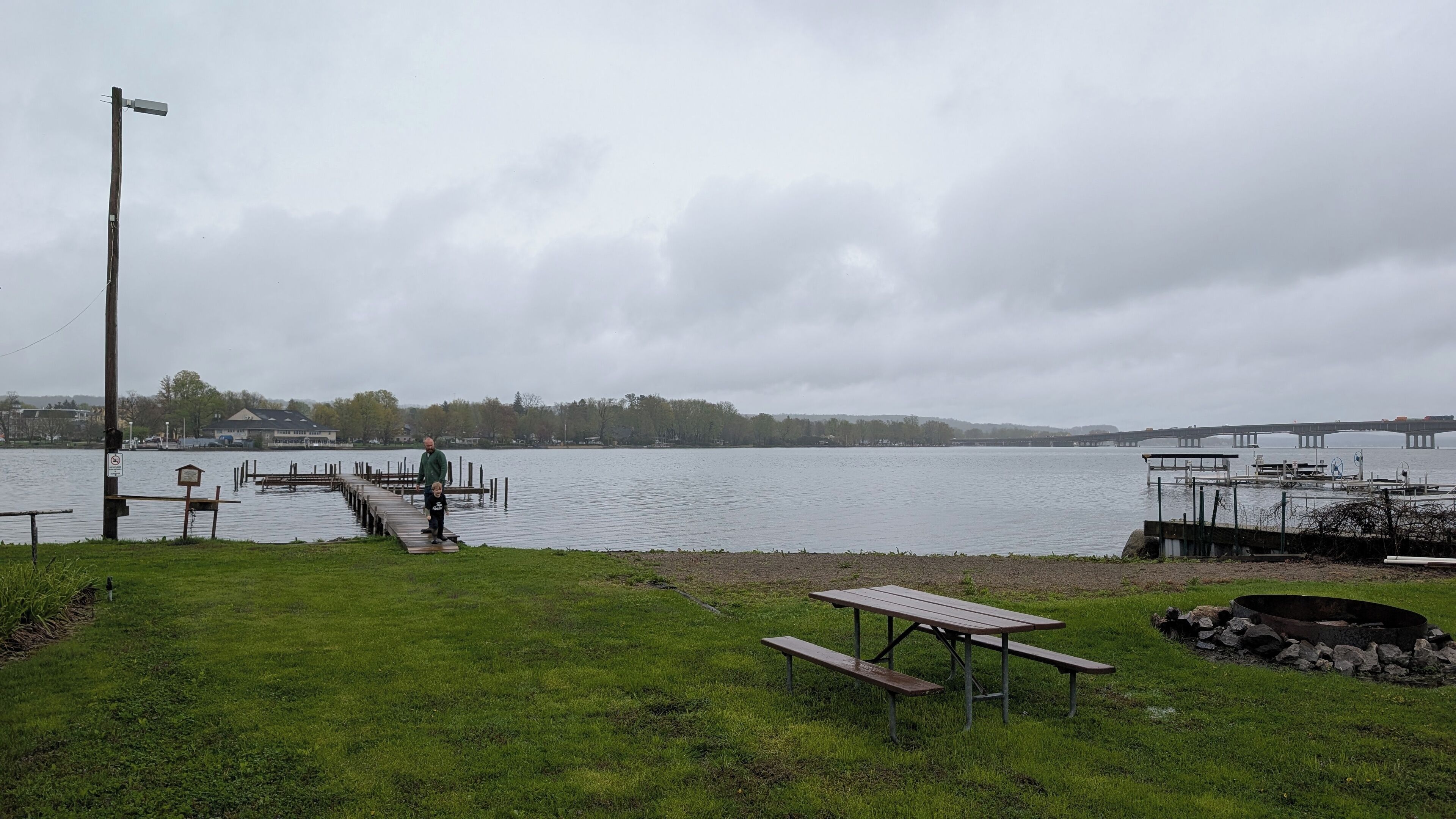 Een park aan het meer met een houten steiger, picknicktafels en een vuurkorf van natuursteen.