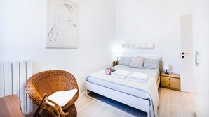 Une chambre à coucher comprenant un lit, une chaise en osier, une table de chevet avec une lampe et une œuvre d’art accrochée au mur.